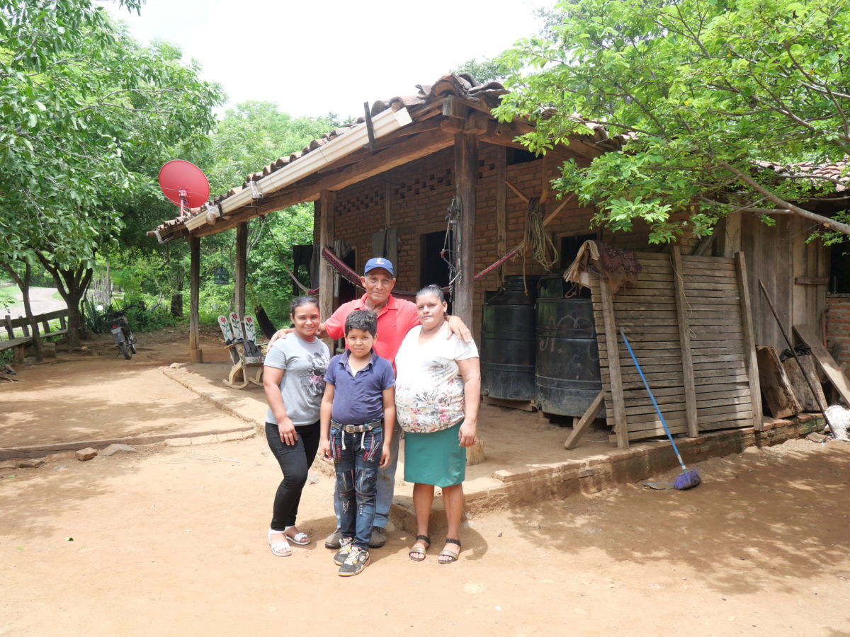 A man wearing a red T-shirt stands outside a wood and brick building with his arms around a woman and a girl. In front of them stands a boy.