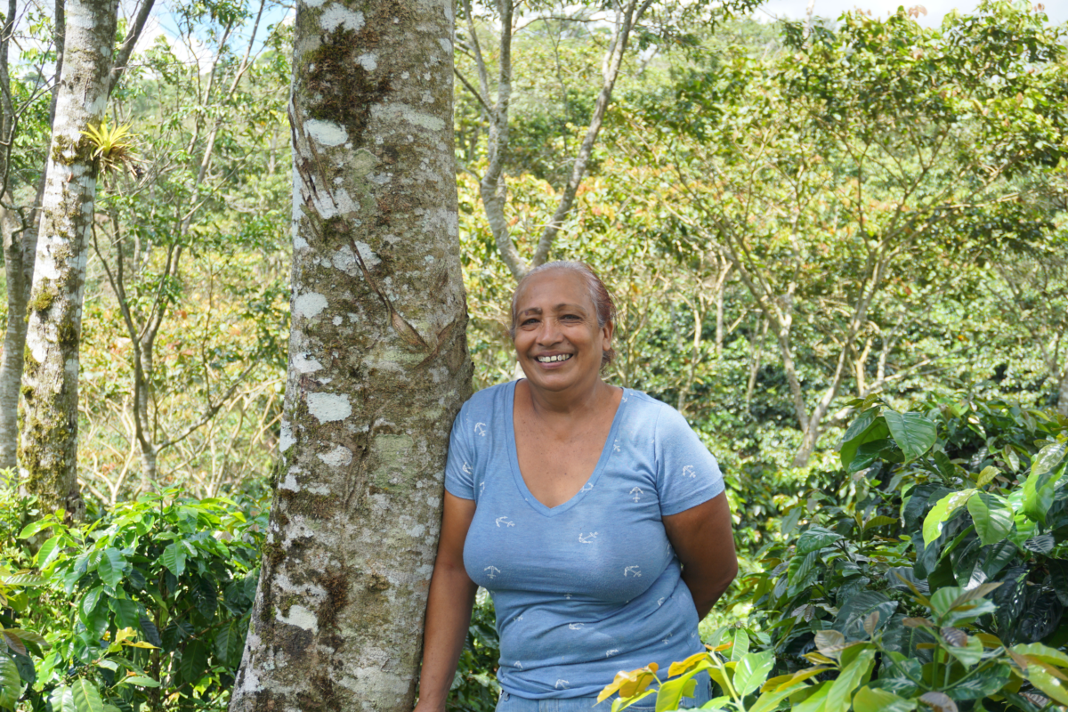 Woman wearing a blue top smiles at the camera as she leans against a tree trunk.